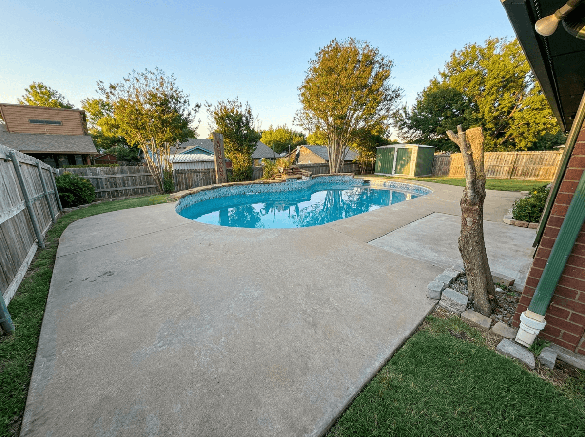 Freeform kidney-shaped pool with wide concrete deck in fenced suburban backyard among mature trees