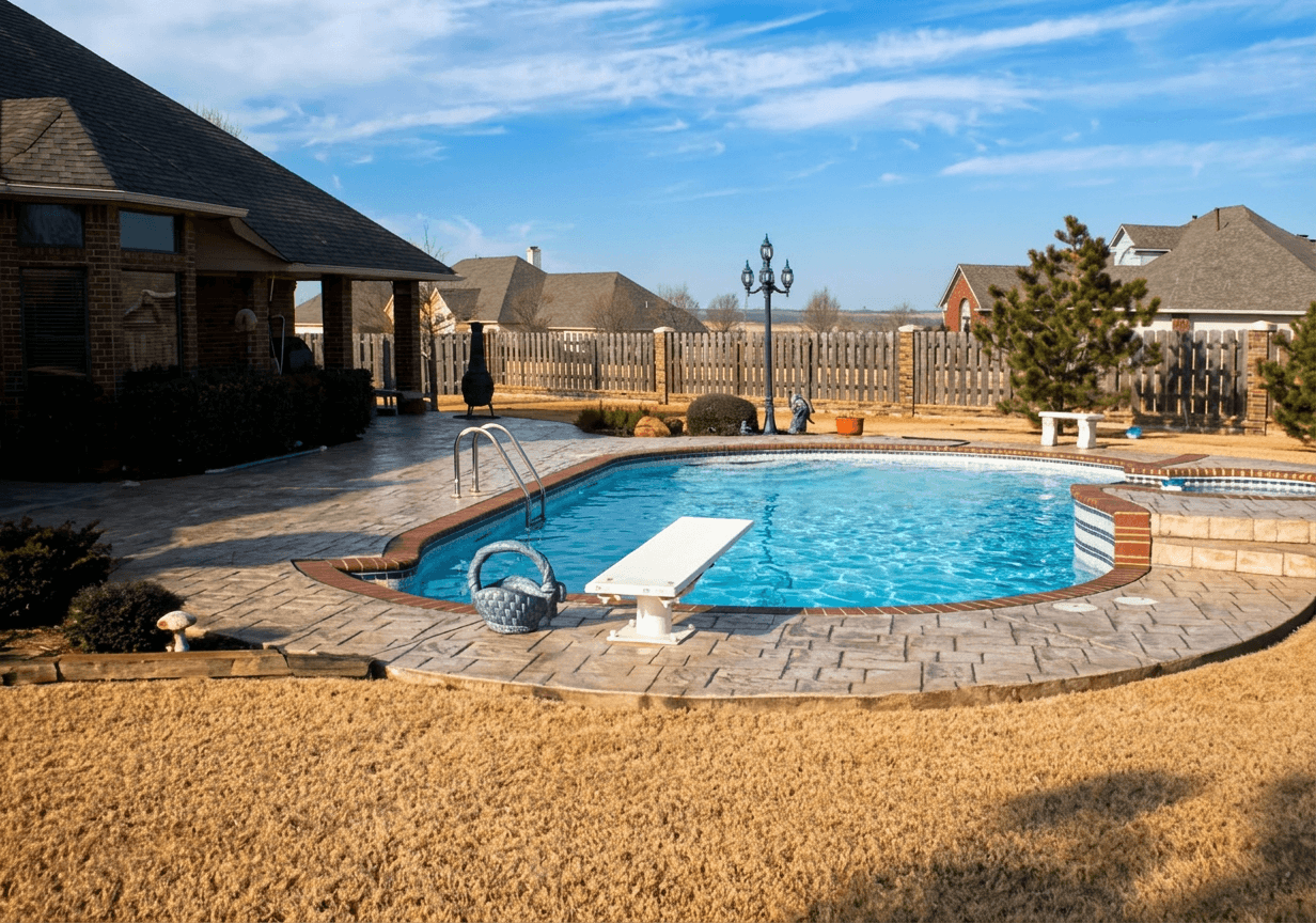 Curved freeform backyard pool with white diving board, tan paver deck and black iron fence under a blue sky