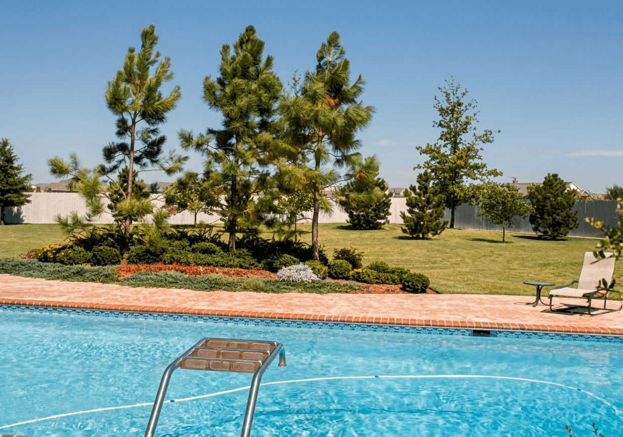 Sunlit pool corner with chrome ladder, brick coping deck, landscaped pines and open green lawn beyond