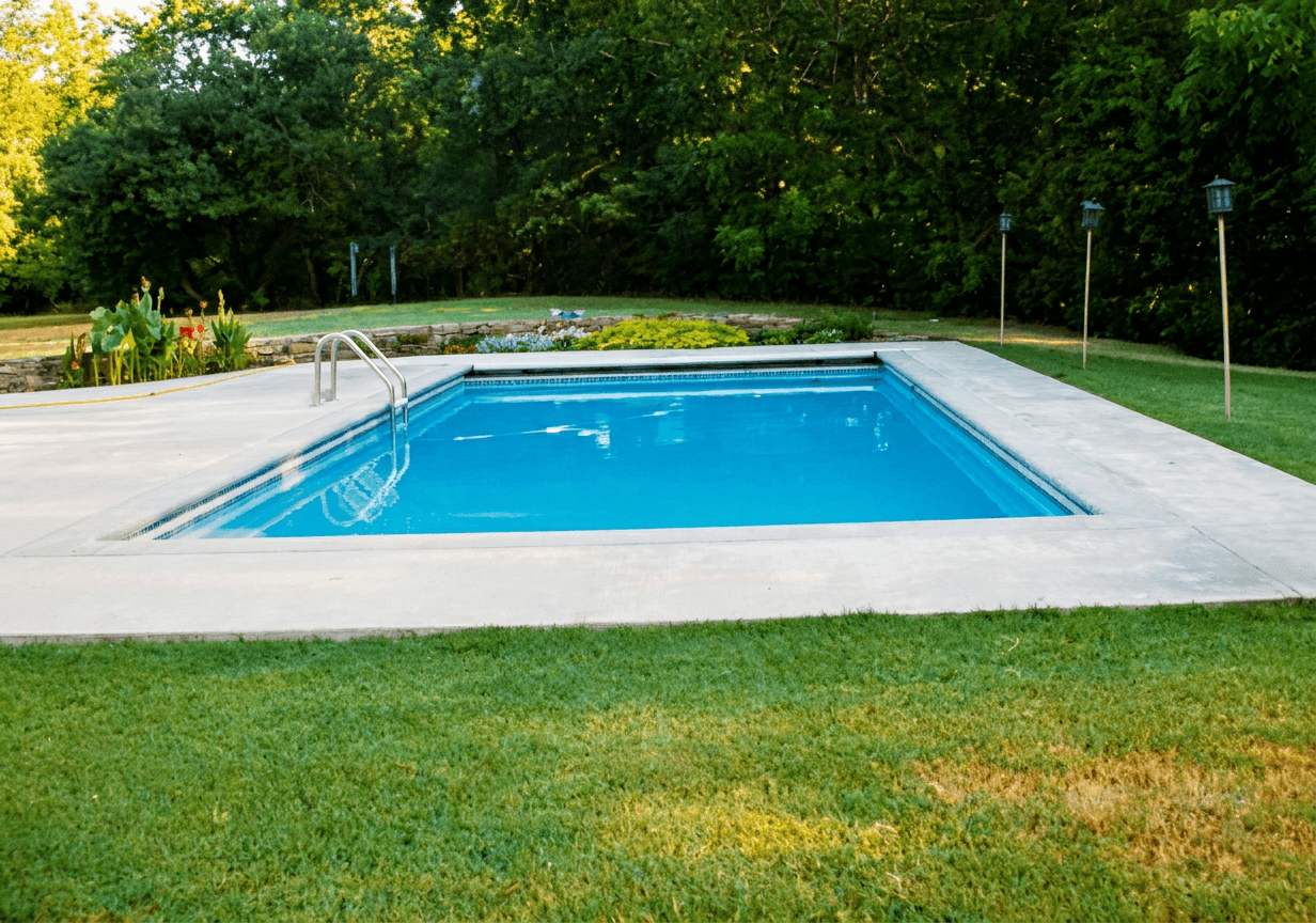 Rectangular in-ground pool with gray concrete deck, corner step handrail, and grassy tree-lined backyard